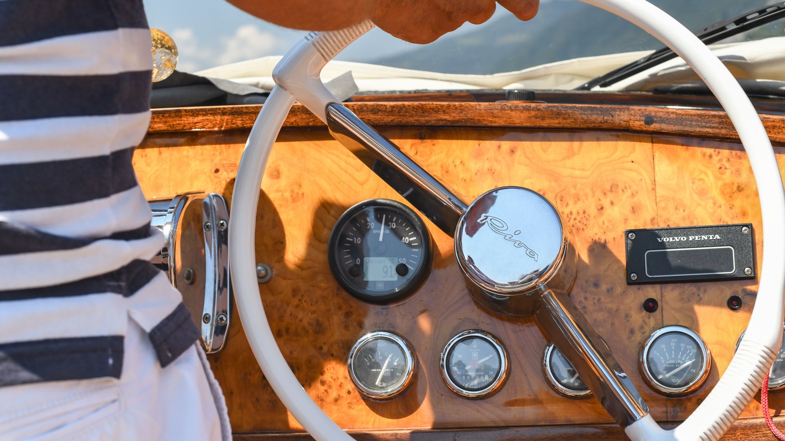 Hand am weißen Steuerrad eines Motorboots mit Holzarmaturenbrett, im Hintergrund ein See mit Bergen