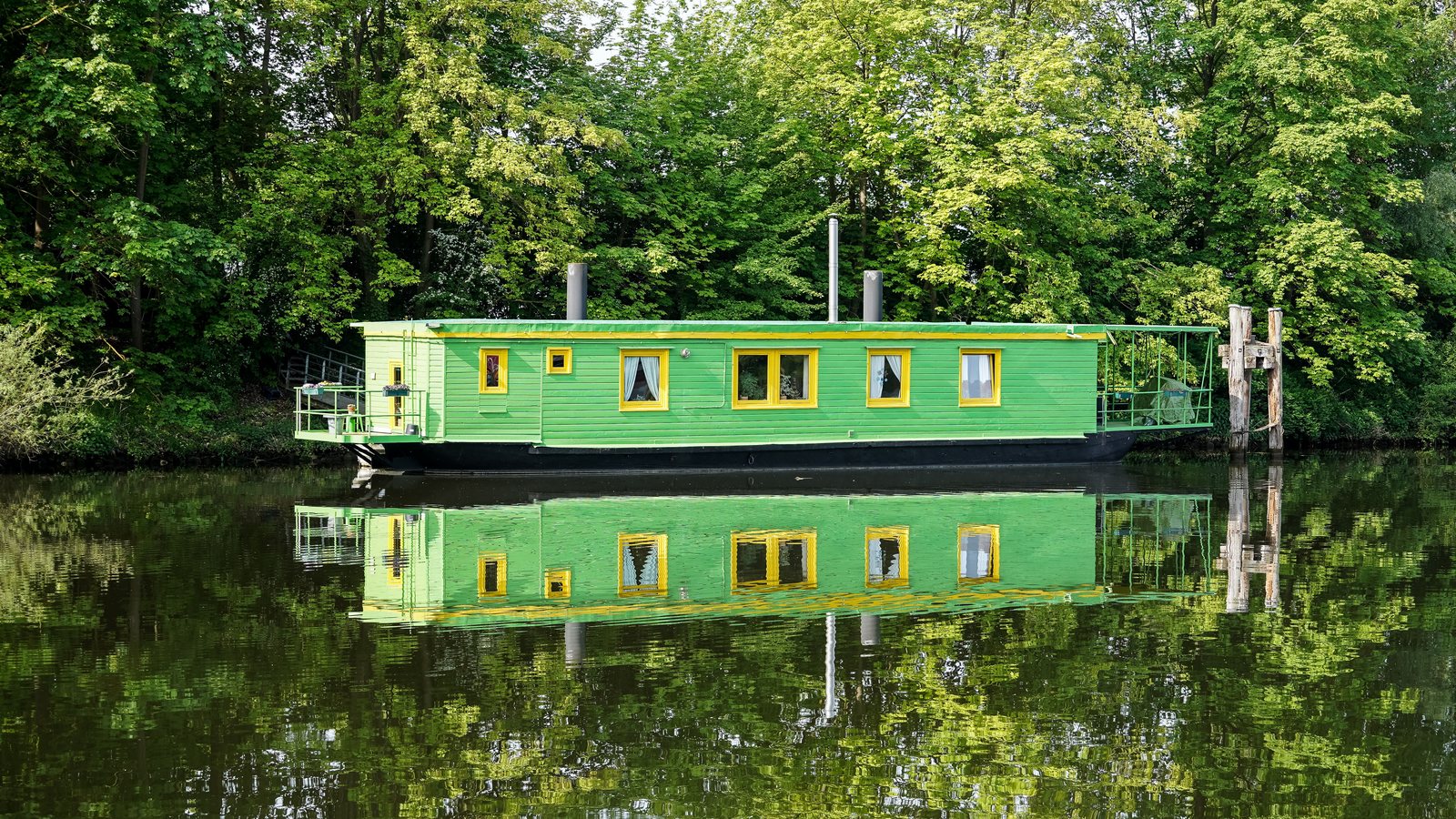 Grünes Hausboot mit gelben Fensterrahmen an einem bewaldeten Kanalufer, das sich im Wasser spiegelt