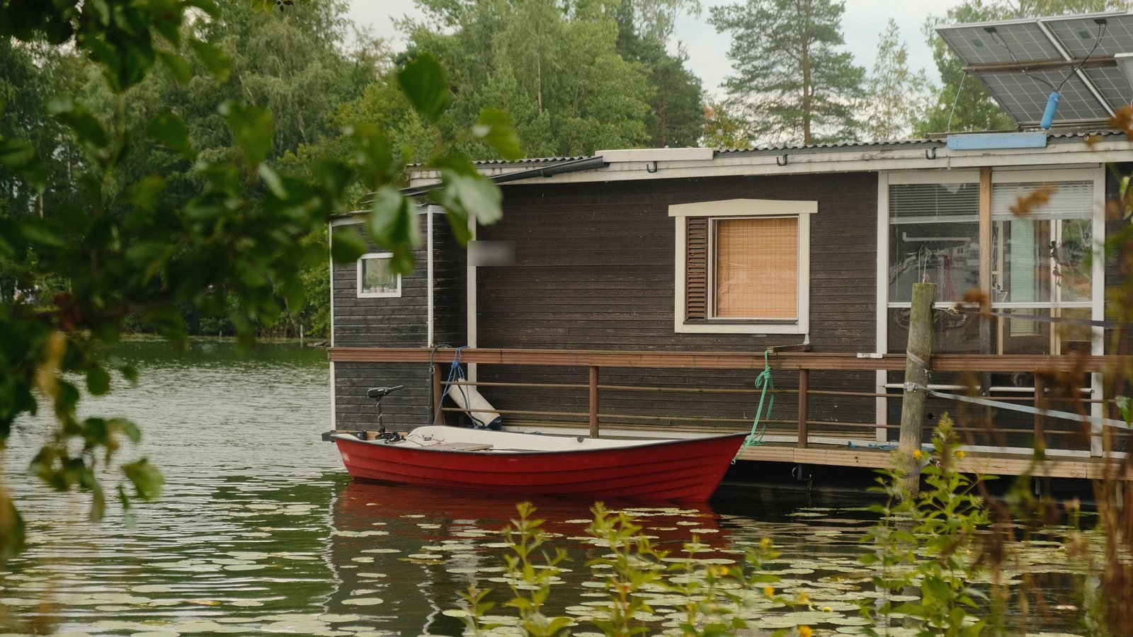 Dunkles Holzhausboot mit Solarmodulen auf dem Dach, davor ein rotes Ruderboot auf einem See